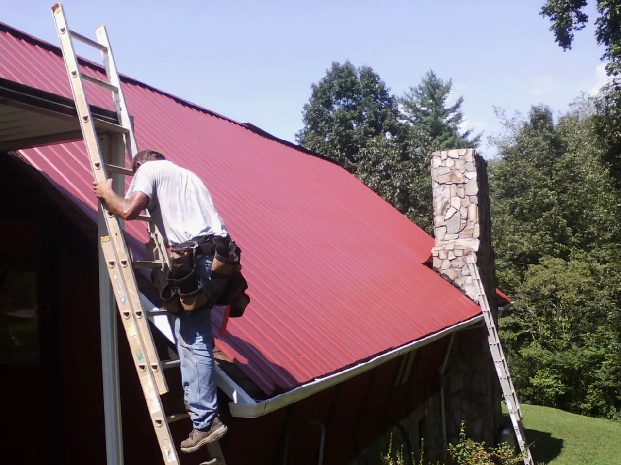 Roofer on a ladder beside a red metal roof and stone chimney — Soffit & Fascia Repair by Roofing Brothers in Coal Valley